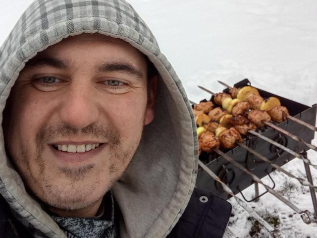 Foto em close-up de DerekPower, um homem de barba usando um capuz xadrez, sorrindo e grelhando espetos de carne em uma churrasqueira de metal. A carne está dourada, com uma grelha ao fundo. Ambiente externo casual..