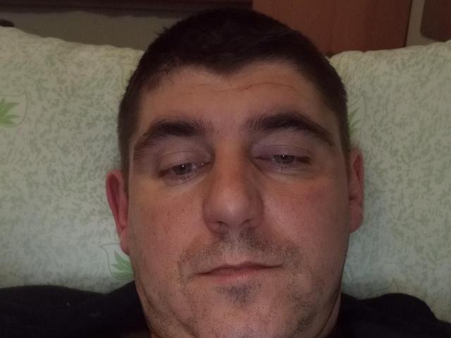 Close-up photo of DerekPower, a middle-aged man with short dark hair, sleepy eyes, and a slight smirk. He lies on a green patterned pillow, his face slightly flushed. Cock visible below.