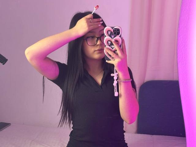 ScarllettDream mit langen, schwarzen Haaren und einer Brille hält ein Telefon mit einer schwarzen Hülle in Herzform und macht ein Selfie. In einem eng anliegenden schwarzen Hemd, stehend in einem schwach beleuchteten Raum..