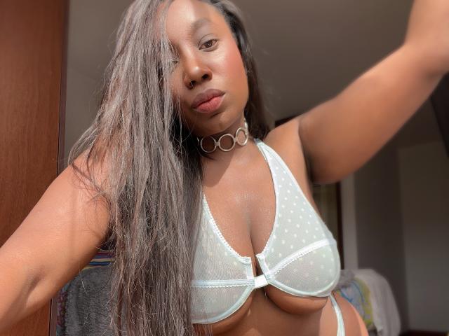 WarisEllen, met nat, lang zwart haar, draagt een witte kanten beha die haar grote borsten accentueert, een choker en een zelfverzekerde pose, recht in de camera kijkend, met zonlicht dat haar huid accentueert..