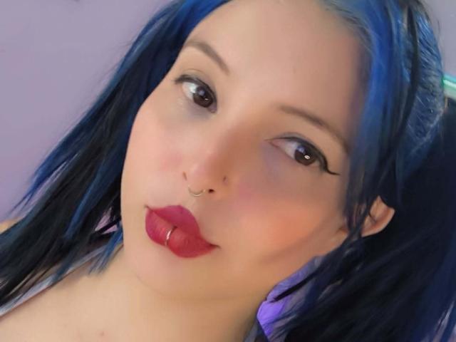 Close-up of SofiaLeal with blue hair, full red lips, and a nose ring. She gazes directly at the camera with a slightly provocative expression, dark eyeliner accentuating her eyes.