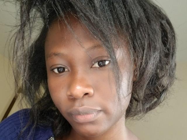 Close-up photo of Blesssing with medium-dark skin, black messy hair, and neutral expression. Blesssing's eyes are slightly squinted, lips slightly parted, showing subtle sexual tension. Wearing dark blue shirt.