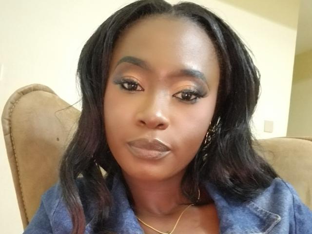 Gros plan de Blesssing, avec une peau foncée, des cheveux noirs et des yeux marron, portant une chemise en jean bleu. Elle a de belles lèvres pulpeuses, rehaussées de gloss, et un sourire subtil qui dégage une séduction sexuelle pleine d'assurance..