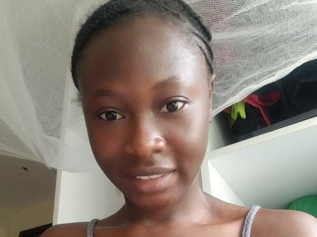 Close-up of Blesssing, dark-skinned woman with braided hair, wearing a gray spaghetti-strap top, looking directly at the camera, slight smile, semi-erotic tension, natural light, white mesh canopy in background.