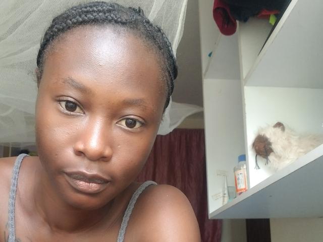 Close-up photograph of Blesssing, a dark-skinned woman with braided hair, glistening skin, and neutral expression. Right side shows a mirror with blurred reflection and toiletries. Sexual tension implied.