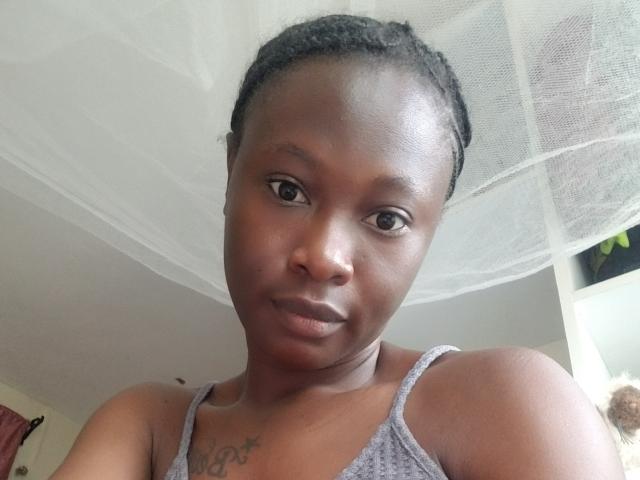 Close-up photograph of Blesssing, a dark-skinned woman with braided hair, wearing a gray tank top, showing a small tattoo on her chest. She looks directly at the camera with a subtle smile. Bright light from the right.