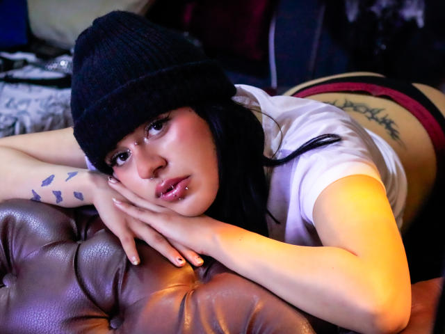 IsadoraFalsone lies face down on a brown leather couch, wearing a black beanie and white shirt, red panties visible, tattoo on her side, tattoos on her forearm, looking seductively at the camera.