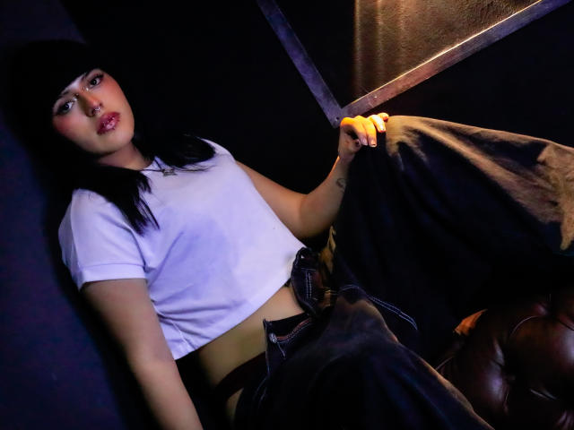 Isadora Falsone se recuesta contra una superficie oscura, sosteniendo una espada brillante en su mano derecha. Ella lleva una camiseta blanca corta y pantalones negros. Su cabello negro tiene flequillo. Mirada increíblemente intensa..