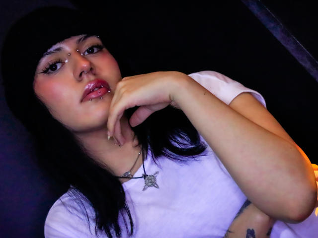 Close-up photo of IsadoraFalsone, Asian woman with black hair, white t-shirt, and nose piercing, hand near mouth, sultry expression, glossy red lips, black background, subtle tattoo on right arm.