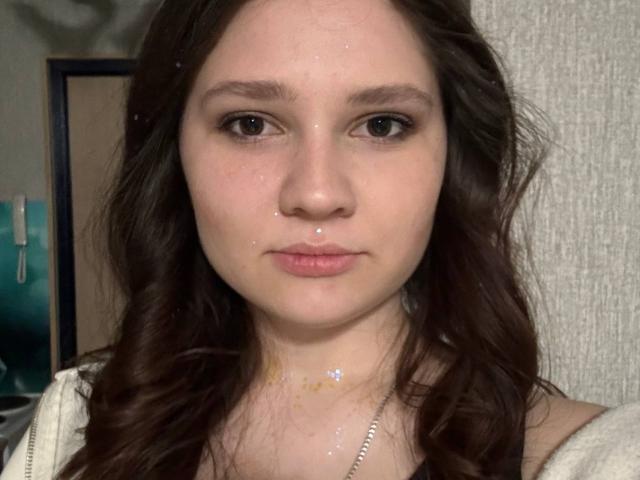 Close-up selfie of CariWannarka, a young woman with fair skin, dark wavy hair, and a septum piercing. She's wearing a white top, looking straight at the camera with a neutral expression.