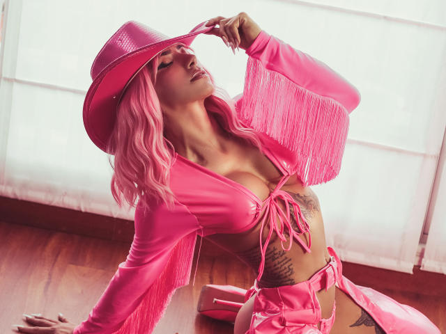 AlessiaFletcher in a pink cowboy hat and matching fringed top, exposing cleavage and tattoos. She's leaning back, hand on hat, glowing white light behind. Vibrant pink, sexy pose, wooden floor.