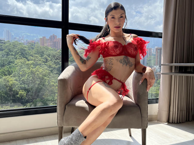 AlessiaFletcher está sentada em uma cadeira cinza, vestindo lingerie vermelha de renda com ombros franzidos e uma fita para meias. Ela tem tatuagens na região das costelas e uma pose confiante, com a luz do sol realçando sua pele. Paisagem urbana ao fundo.