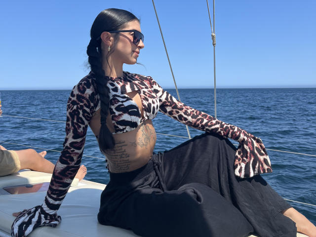 Alessia Fletcher, met zwart haar, draagt een top met een zebraprint, zwarte broek en een zonnebril, en zit op een boot, waarbij ze haar getatoeerde buik. Achtergrond met een blauwe oceaan. De haan is niet zichtbaar. Er is een lichte seksuele spanning..
