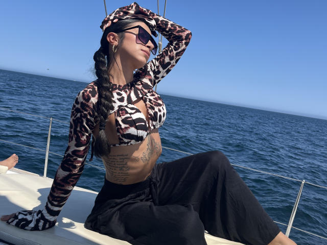 Alessia Fletcher, gekleed in een zwart-wit gestreept topje en zwarte broek, zit op een boot, met haar hand op haar hoofd, en heeft tatoeages. zichtbaar, blauwe oceaan op de achtergrond, heldere lucht, verlicht door de zon, sexy pose.