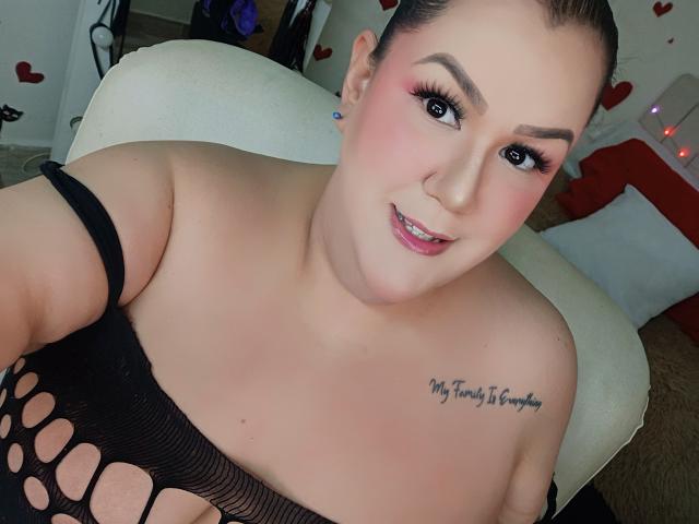 Close-up of GiiannaMorrys with dark hair, pink blush, and black fishnet top showing cleavage. Tattoo on right shoulder reads "My Tony S. Favorite." Sexual tension, direct gaze, confident smile.
