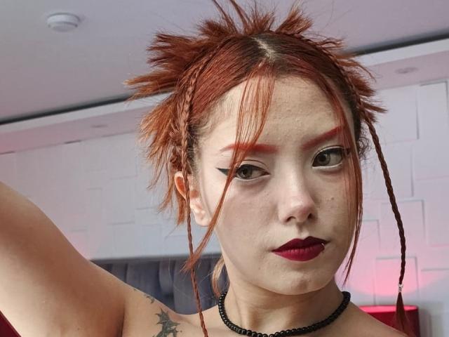 Primer plano de EmmaThomsonn con cabello rojo despeinado, lápiz labial rojo oscuro, delineador de ojos negro y un collar negro. Ella levanta su brazo, mostrando un tatuaje de una estrella en su hombro. Tensión sexual, mirada directa..