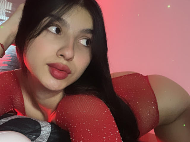 MilanMoon with dark straight hair, red lipstick, and a red sparkly top, leaning forward, looking to the side with a flirtatious expression, red lighting in the background. Slight sexual tension.