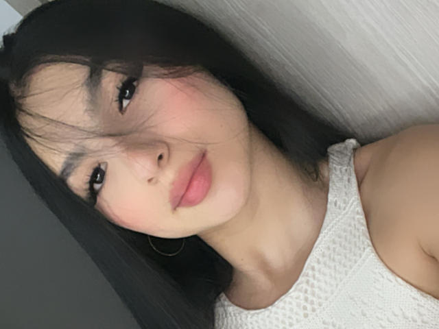 Close-up of MilanMoon, an Asian woman with black hair and bangs, wearing a white lace top, looking at the camera with slightly parted pink lips. Her expression is seductive and relaxed.