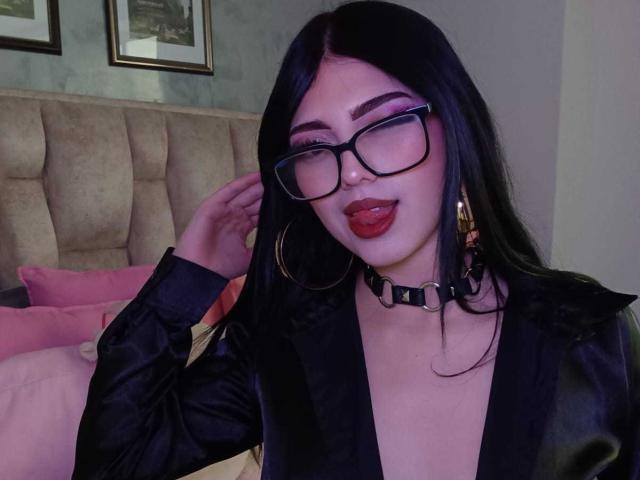 Isabella Rossi, una mujer de cabello negro, con gafas, lápiz labial rojo, lengua afuera, collar negro, blusa negra, aretes de aro dorados, mano en el pelo, dormitorio, cabecero acolchado, almohadas rosas, cuadros enmarcados en la pared. Increíblemente atractivo/a..