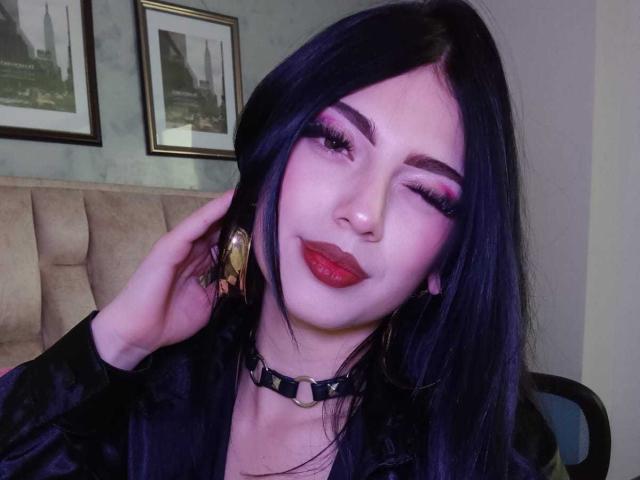 Photograph of IsabellaaRossi, fair-skinned with black hair, heavy pink eyeshadow, red lipstick, and a black choker. She's winking, touching her ear with a hand, looking sexy. Framed pictures in the background.
