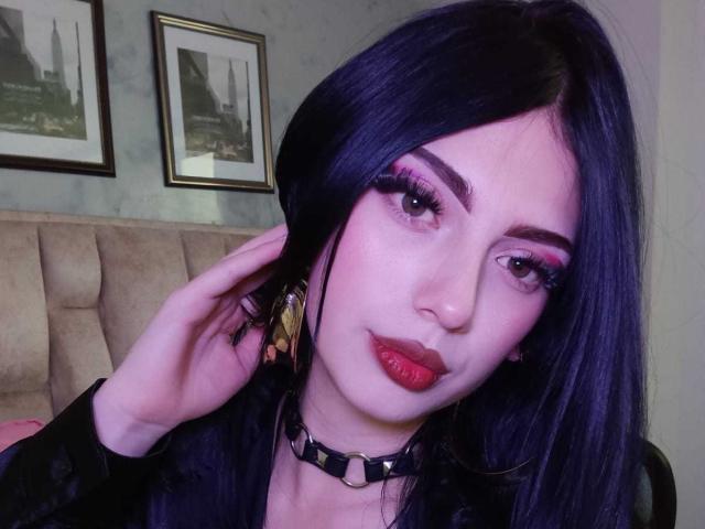 Selfie de perto de Isabella Rossi, com pele clara, cabelo preto, batom vermelho, maquiagem pesada nos olhos, colar preto e dourado. brincos. Ela toca sua orelha, com uma obra de arte que retrata uma paisagem urbana ao fundo. Muito quente.