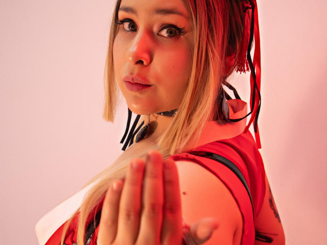 Nahaufnahme von DandelionQueen mit roten Highlights in ihrem Haar, die über ihre Schulter schaut und einen verführerischen Ausdruck hat. Ihre Hand ist vor ihrer Brust erhoben und betont so ihre Rundungen. Helle Rot- und Weißtöne dominieren..