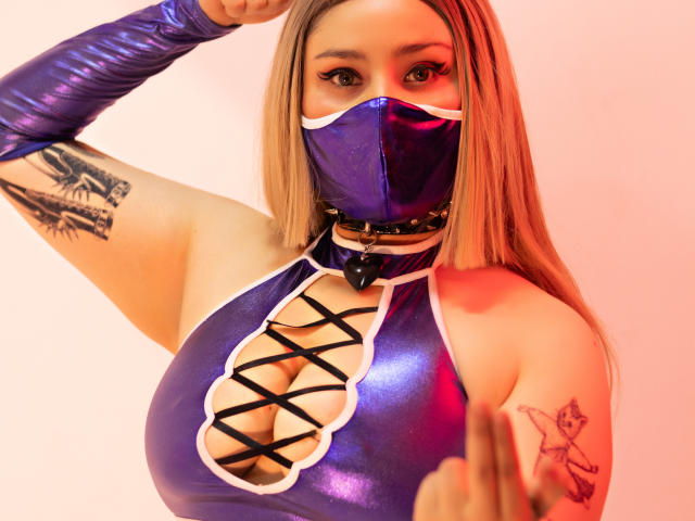 Foto ravvicinata di DandelionQueen, una donna formosa con grandi seni, che indossa una tuta intera blu in lattice lucido con motivo a croce. lacci, una mascherina nera e guanti viola per le braccia. Braccio tatuato sollevato. Incredibilmente sexy..