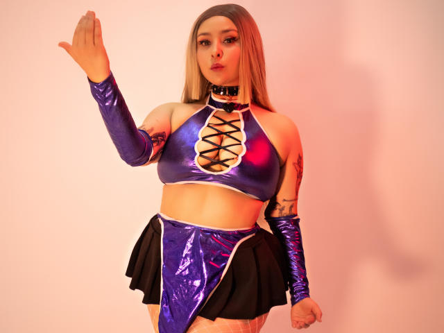 DandelionQueen, com longos cabelos loiros, vestindo uma blusa cropped azul e preta brilhante com cadarços, munhequeiras combinando e uma saia curta. Saia preta.
ela levanta uma mão, com uma expressão intensa. A iluminação neon.