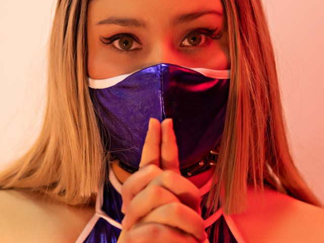 Close-up of DandelionQueen with long blonde hair, wearing a blue face mask, covering her mouth with her fingers, red light illuminating her eyes, intense gaze, serious expression, bold colors.