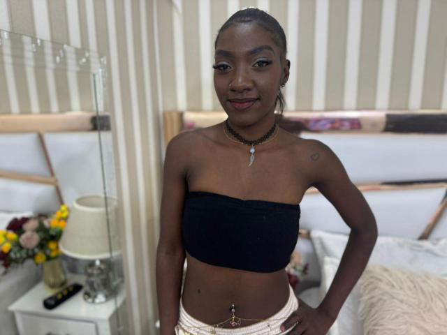 Photographie de MiaPeints debout dans une chambre. Elle porte un haut noir sans bretelles, un bas en dentelle blanche et un collier ras du cou avec un pendentif. Elle a la peau foncée, les cheveux tressés et un sourire confiant. Son nombril est percé. Lit avec des draps blancs et des fleurs jaunes en arrière-plan..