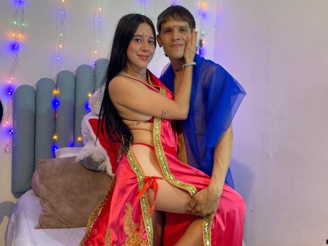 AnjaliRohan, in a red and gold outfit, sits on a bed with a guy in a blue sheer shirt. She's straddling him, her hand on his face, string lights in background.