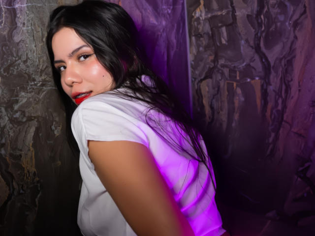 Fotografia de MaraCortes, virada para longe, mas olhando por cima do ombro com uma expressão sedutora. Cabelo preto, batom vermelho, camisa branca. Parede escura com textura ao fundo, com luz roxa. Muito atraente..