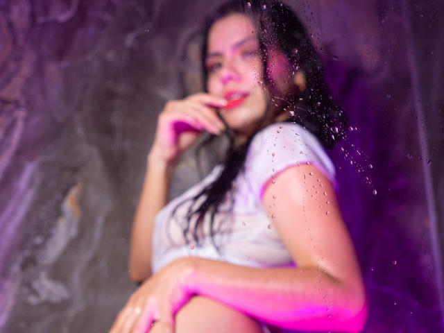 Foto borrada de MaraCortes com cabelo escuro e molhado, top branco, iluminação rosa e roxa, dedo perto da boca, parede molhada. fundo, sorriso discreto, sensualidade, pose sugestiva, gotas de água.