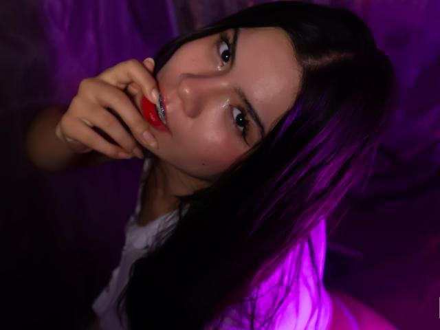 Close-up photograph of MaraCortes with dark hair, biting red lipstick, gazing seductively into the camera. Purple neon light illuminates her face, creating intense sexual tension. Her hand rests near her mouth.