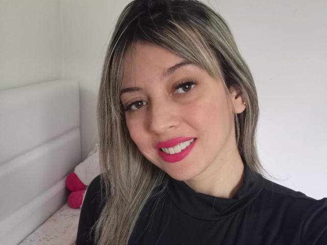 Selfie de perto de Lancatelli, com cabelo castanho claro, batom rosa e camisa preta. Lancatelli tem um sorriso adorável, olhos castanhos e uma pequena perfuração no nariz. Fundo branco com um travesseiro rosa.