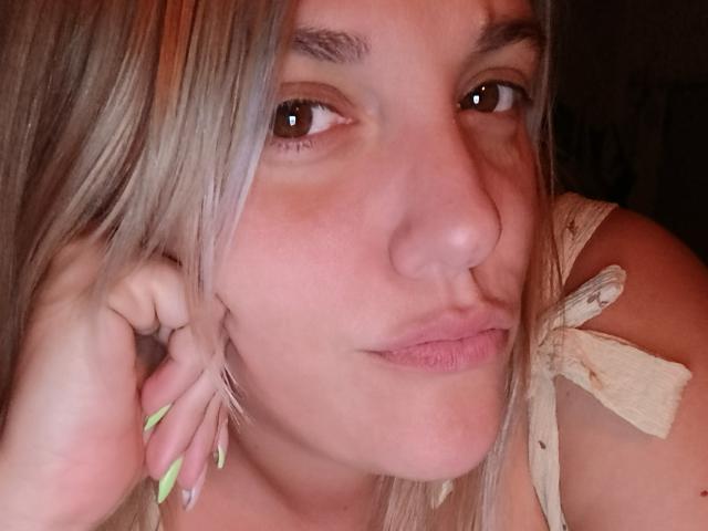 Foto ravvicinata di CurvyCami, capelli biondi, occhi marroni, labbra piene, unghie verdi neon, nastro beige sulla spalla, leggermente pelle arrossata, sguardo intenso, sottile tensione sessuale, contatto visivo diretto.