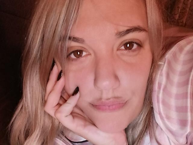 Nahaufnahme-Selfie von CurvyCami, hellblondes Haar, braune Augen, leichtes Lächeln, Hand berührt die Wange, schwarzer Nagellack. Ein rosa und weiß gestreiftes Hemd, sanftes rosafarbenes Licht, lässig, intime Atmosphäre..