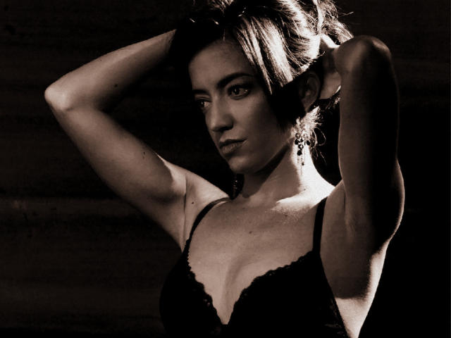 Fotografia in bianco e nero di CallMeCarlota, una donna attraente con capelli scuri, che indossa una camicetta di pizzo scollata, con le braccia alzate e le mani dietro la testa, sfondo scuro, sguardo intenso, orecchino, alto contrasto.