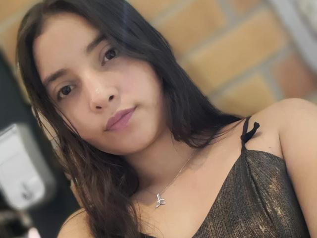 Tiffany Collins, com longos cabelos pretos e lisos, vestindo uma blusa preta brilhante com alças finas, olhando diretamente para o câmera com uma expressão neutra, usando um colar de prata com um pingente de asa..