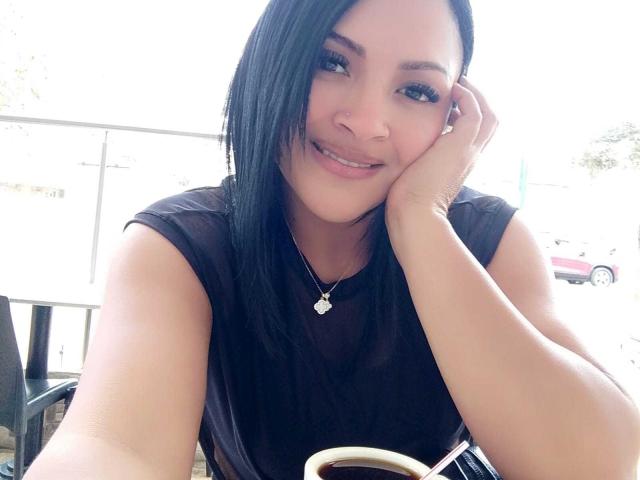 Fotografie von EstrellaSmmith mit glattem, schwarzem Haar, die lächelnd in die Kamera schaut und ihre Wange in ihrer Hand ruht. Sie trägt ein schwarzes Top ohne Ärmel und eine silberne Halskette. Im Hintergrund ist eine helle Landschaft zu sehen. Leichte sexuelle Anspannung.