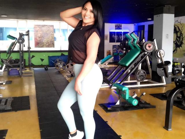 Fotografia de EstrellaSmmith em uma academia, em pé com uma mão atrás da cabeça, vestindo uma blusa preta sem mangas e calças brancas. calças, ao lado das máquinas de musculação, exibindo uma pose sexy e confiante..