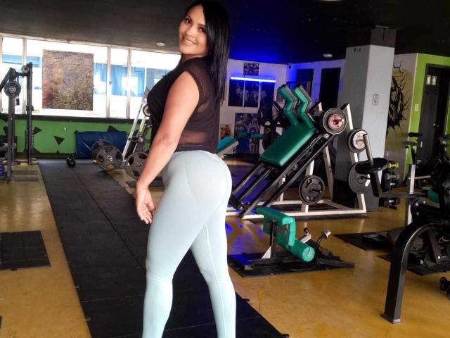 Foto van EstrellaSmmith in een sportschool, staand met de rug naar de camera, kijkend over de schouder, zwarte top, strakke witte broek. leggings, die haar billen accentueren, sportschoolapparatuur op de achtergrond, blauwe verlichting.