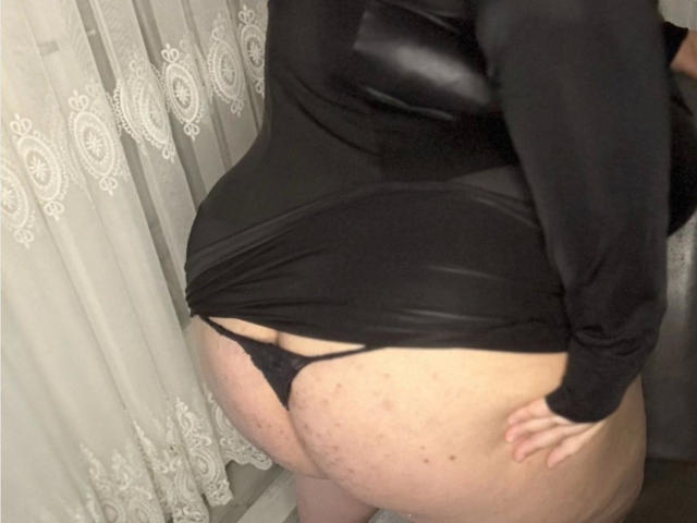 Curvy-jasmin1990-hot stands with their back to the camera, black top pulled up, exposing their round, pale buttocks in black thong. White lace curtain in background. Light source from top.