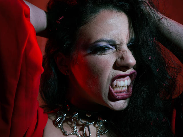 Close-up of MiniBeast with dark, wavy hair, eyes closed, mouth open in a snarl, tongue visible, wearing a metallic chain collar, red fabric in the background, intense expression, dramatic lighting.