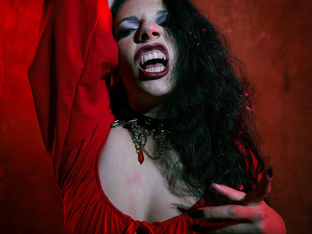 Close-up van MiniBeast met lang, krullend zwart haar, een rode jurk en opvallige make-up. Ze bijt op haar lip, haar arm is omhoog, er is rood licht, ze heeft een intense uitdrukking, en ze draagt een ketting met een rode hanger. Heel aantrekkelijk..