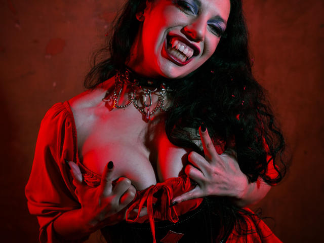 MiniBeast with dark, wavy hair, red-lit face, and heavy makeup, smiling widely, hands pressing against red dress, exposing large breasts, wearing a chunky necklace, intense sexual tension.