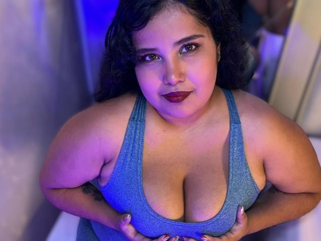 Cassandra Valmont, una mujer de talla grande con cabello oscuro, vestida con una camiseta azul brillante, con grandes senos, con las manos tirando hacia abajo. escote, iluminación morada y azul, mirada seductora, labial oscuro, expresión de confianza..