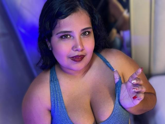 Cassandra Valmont, eine übergewichtige Frau mit dunklem Haar, trägt ein glitzerndes blaues Tanktop mit tiefem Ausschnitt, rotem Lippenstift und hält... Eine gepflegte Hand mit weißen und roten Nägeln, blaues Licht, suggestiver Blick..