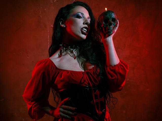 MiniBeast avec des cheveux noirs et mouillés, une robe rouge à une épaule, tenant un crâne noir, lumière rouge, expression intense, peau mouillée. collier noir, éclat rouge, pose suggestive, tension érotique, ambiance gothique..