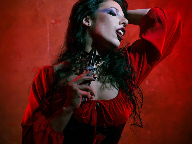 MiniBeast met lang, nat zwart haar, gekleed in een rode jurk, hand achter het hoofd, andere hand op de borst, rood licht, open mond, ketting, rode achtergrond, intense uitdrukking, seksuele spanning.